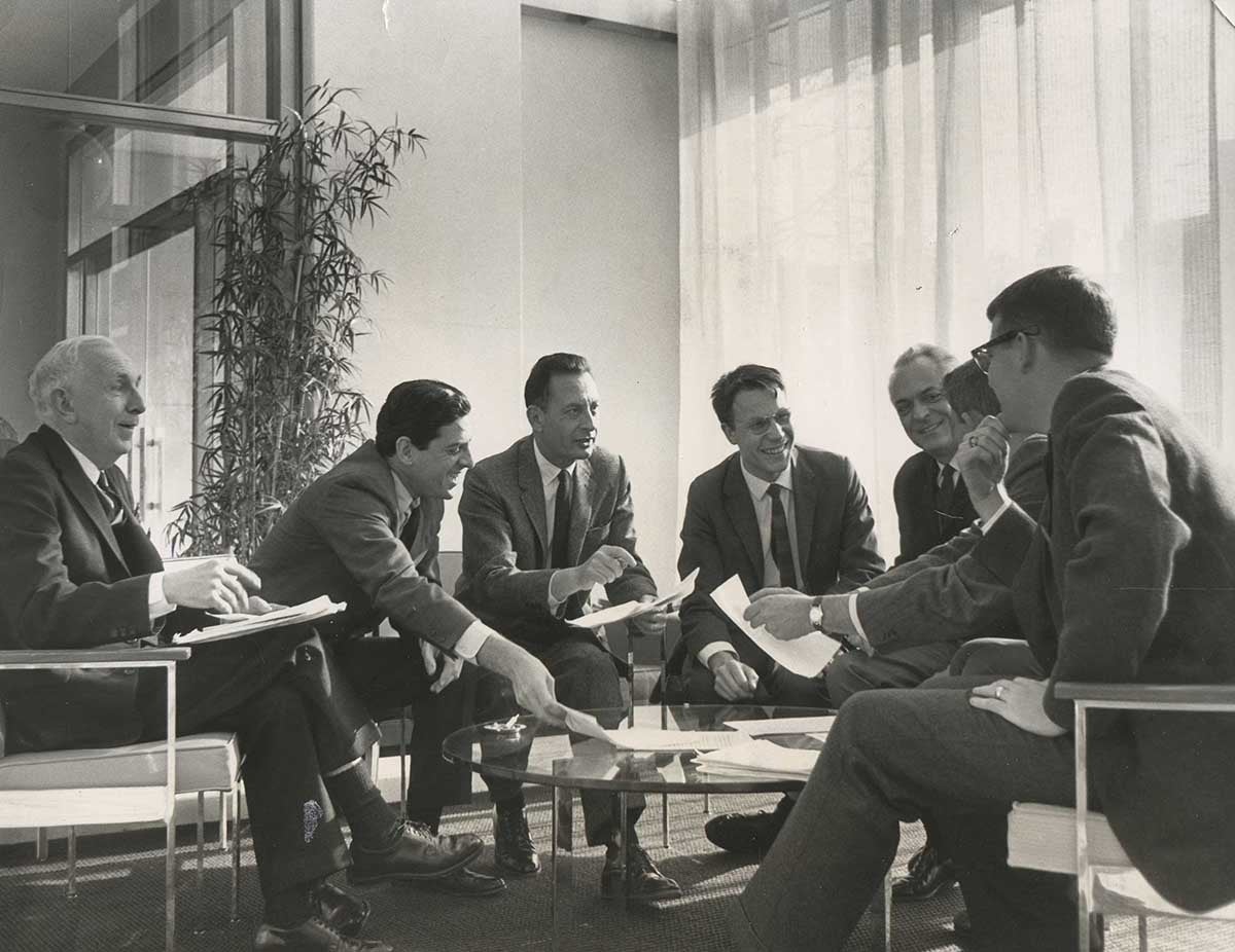 A group of men sitting around a table wearing suits with light streaming in the curtain behind them