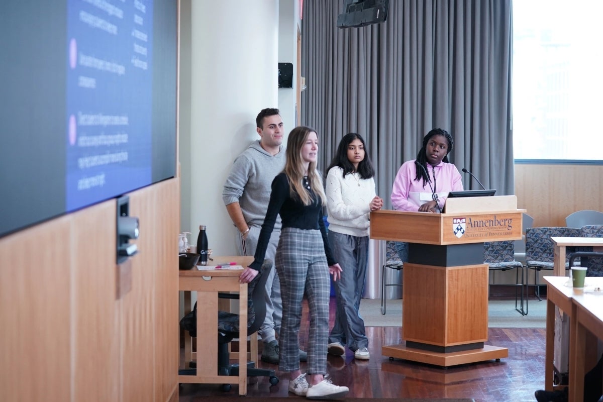 Four students stand in front of a podium. To their right, a screen shows part of a presentation slide