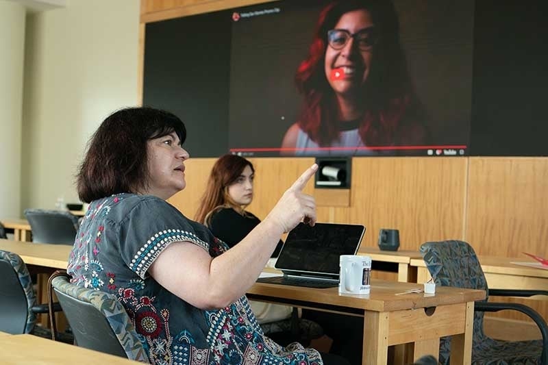 Mihaela Popescu points as she speaks. A woman sits in the background, and a video with a woman's face is on a large TV screen