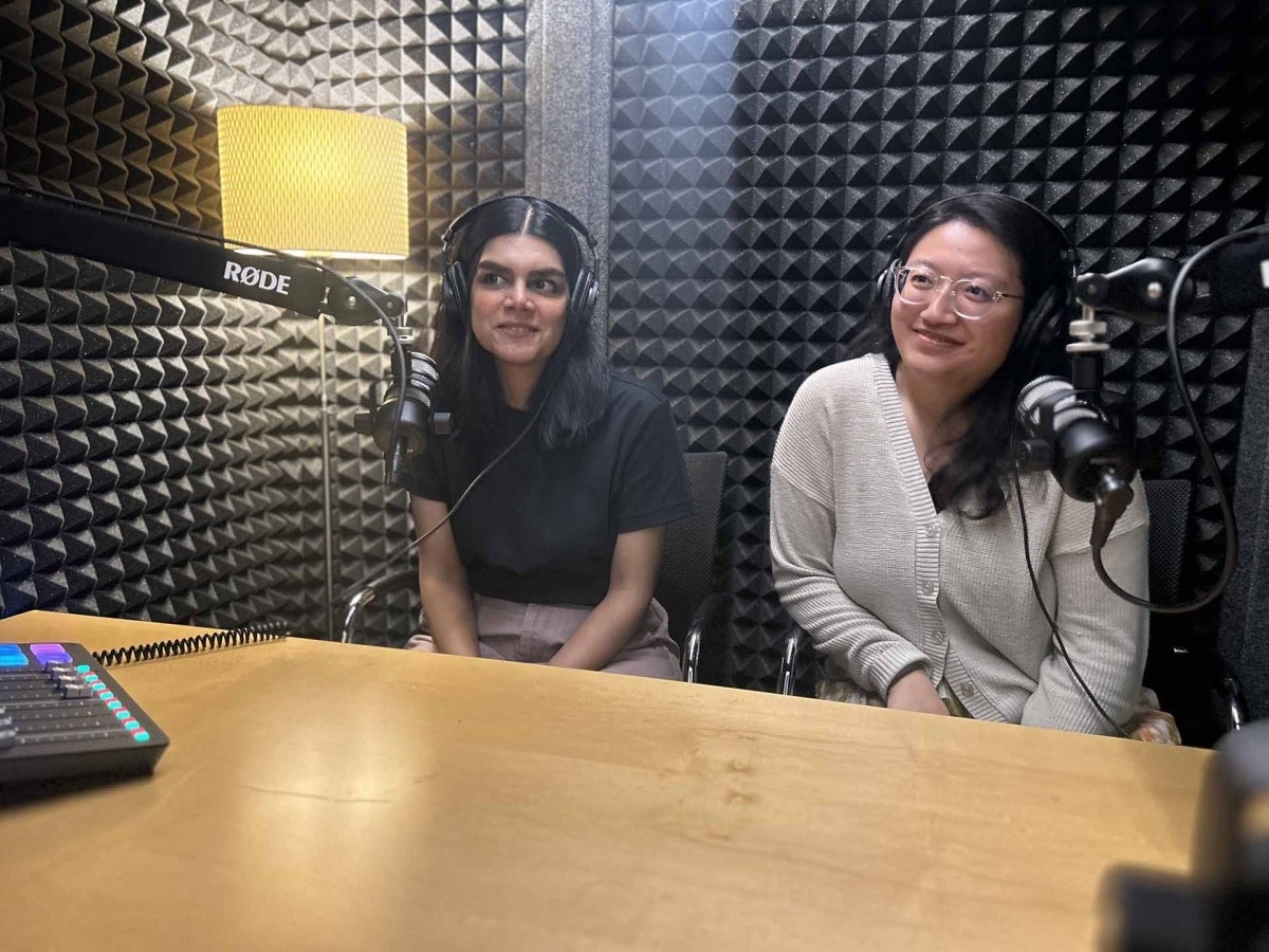 Two people sit in front of microphones at a table in a room with textured foam on the walls