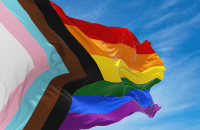 A Progress Pride Flag blows in the wind against a blue sky with several clouds