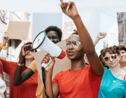 MIC_PME_700x550 Photo Credit Rawpixel on iStock.png Woman in an orange shirt at protest with a megaphone, Photo Credit: Rawpixel on iStock