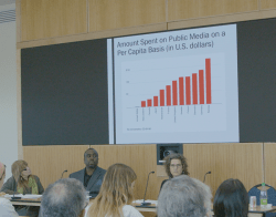 Pickard_Still_5.png People seated at a table facing an audience, in front of a slide with the headline "Amount spent on public media on a per capita basis (in US dollars)"