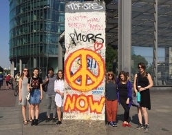 Summerculture Berlin 700 x 550.jpg Seven people standing next to a chunk of the Berlin Wall with a graffiti peace sign and the word "now"