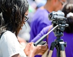 MIC Carnegie Fellowship 700x550 Photo credit anouchka on iStock.jpg Woman standing in front of a camera and looking at her cell phone at an outdoor event, Photo Credit anouchka on iStock