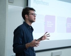 Student Speaking 700 x 550.jpg Student standing up in front of a screen and speaking. Slide is blurry, but headline reads "Hypotheses"