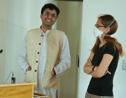 Prateekshit and Emily 700 x 550.jpg Person stands behind a podium smiling, with another person wearing a mask with arms folded looking at them