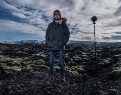 VR Greenland Zane 700 x 550.jpg Person in winter gear stands outdoors in an eerie-feeling landscape with rocks and plants and dramatic clouds in the sky. A VR camera on a tripod also stands in the foreground