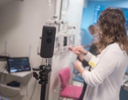 VR Nursing 700 x 550.jpg A camera on a tripod is in the foreground with medical people doing unspecified tasks indoors behind it