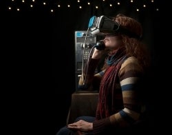 VR Payphone 700 x 550.jpg Person seated in a dark room with a VR headset on, listening to the receiver of a payphone which sits behind them