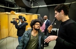 Student holds microphone up to second student. Woman in background is filming something.