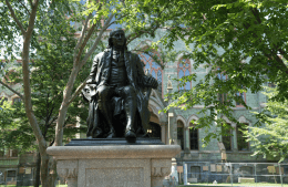 Ben Franklin statue with College Hall, a stone building, and trees in the background 