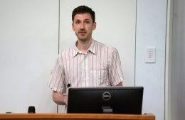 Lee McGuigan stands behind a wooden desk giving a speech. He is wearing a green, purple, pink and ivory vertically-striped button-down shirt. There is a black Dell monitor on the desk. 