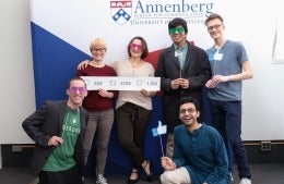 6 attendees of the sixth graduate symposium pose in front of a red, white and blue background with the Annenberg School for Communication logo at the top. 4 of them are standing. The left two hold a prop that has the twitter comment icon and the number 26k, the retweet icon with 328k, and the like icon with 1.6m. The middle two wear red and green sunglasses. The right-most person standing and the one kneeling hold a prop of the Facebook like prop. The left-kneeler wears purple shades and a bowtie prop 
