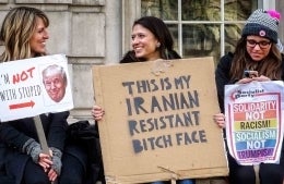 Three women seated while holding signs. The two to the left are smiling at each other, and the left-most one's white sign says " Im not with stupid", with 'not' in red, capitalised letters and has a red arrow pointed to a cut-out of Donald Trump's face. The one in the middle has a cardboard sign which reads "This is my Iranian Resistant Bitch Face". Finally, the lady to the right, who is typing on her phone, is holding a sign that reads "Solidarity not Racism! Socialism not Trumpism" on a rainbow background