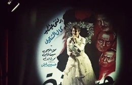 Singer wearing a white dress on a stage being spotlighted. The illuminated background has Arabic written in blue and black and faces of four men in red.