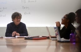 Joan Garry seated at the head of a table in front of a white board. She is seated with students, and is laughing with one to her left. 