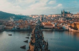 City of Prague. There is a body of water in the foreground parted by a bridge filled with people, and the background is filled with buildings of various heights and widths; photo credit: Anthony Delanoix / Unsplash