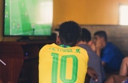 Back shot of someone wearing a Neymar JR Brazilian football jersey while watching the television that has a football match going on. Other people watching the show are in the background of the image. photo credit Gustavo Ferreira / Unsplash