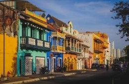 Street-view of a line of houses painted in colors such as turquoise and dark green, blue and yellow, and orange and yellow and white. All of the houses have a balcony; photo credit: Leandro Loureiro / Unsplash