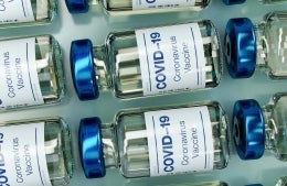 Blue-capped vials of COVID-19 vaccines lined up. The vials are glass, and they have a white label with the words 'COVID-19 Coronavirus Vaccine' in blue, photo credit Daniel Schludi / Unsplash