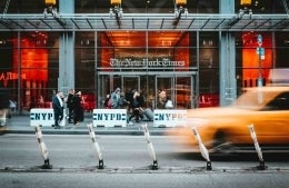 Front of The New York Times building in New York City. There are people on the sidewalk walking in front of it, and on the road is a yellow taxi driving by. photo credit Stéphan Valentin/Unsplash