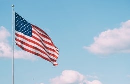 American flag fluttering in the wind on a bright day with clouds in the background, photo credit Aaron Burden / Unsplash