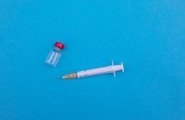 needle and vaccine bottle laying on a blue surface, photo credit Markus Spiske / Unsplash