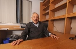 Elihu Katz smiling seated behind an empty desk with his hands on the desk.