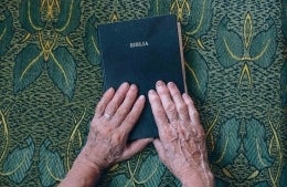 Hands of an old person on the cover of a bible labeled "Biblia" and on an ornate green tapestry