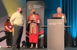 Bob Hornik standing with a person holding a plaque and a person reading from a podium reading "Palais des Congres de Paris." An International Communication Association logo is also visible.