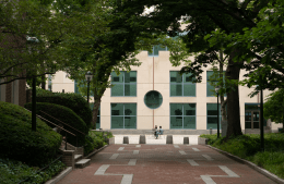 Brick walkway lined with trees that leads to a large plaza and building. Two people sit at a table on the plaza.