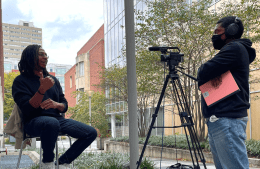 Antoine-Header.png Outside, one person sits on a stool in front of a camera while being interviewed by a person standing behind a camera