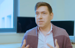 Juan Llamas-Rodriguez sitting in front of two computer screens and speaking