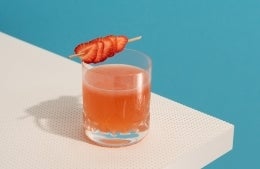 Drinking glass with red/orange colored beverage garnished with a strawberry skewer, sitting on white table against solid blue background