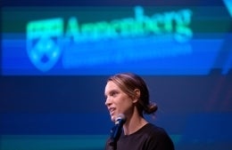 Lauren Hitt speaking at a microphone in front of a large screen with the Annenberg logo blurred in the background