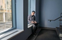 A man sitting on a window ledge in a stairwell, looking up from his laptop