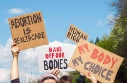 Hands holding protest signs reading: Abortion is Healthcare, Bans off our bodies, My Body My Choice, and Human Rights