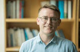 t: David Lydon-Staley, wearing round glasses and a light blue button-up shirt, smiling in front of a wooden bookshelf filled with books.