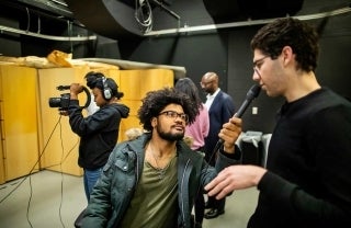 Student holds microphone up to second student. Woman in background is filming something.