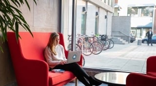 Student on Red Couch FWF.jpg Woman seated on red couch with a laptop in front of a window. A bike rack is visible through the window.