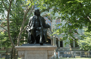 Ben Franklin statue with College Hall, a stone building, and trees in the background