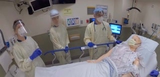 Three clinicians in full PPE in a hospital room standing around a medical dummy in a bed