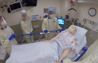 Three clinicians in full PPE in a hospital room standing around a medical dummy in a bed