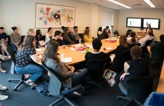 Large group of people sitting around a conference table and along the walls, as a woman presents