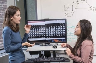 Photo of Emily Falk and Elisa Baek speaking in front of a computer with brain image scans
