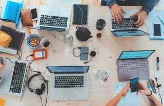 View from above of people sitting at a table with laptops and other miscellaneous objects, photo credit Marvin Meyer / Unsplash