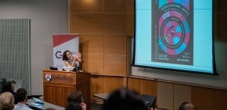 Person speaking at an Annenberg Podium, gesturing at a screen that has a poster with the headline "Secrets Without Agents: From Big Brother to Big Data"
