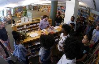 Man in a purple shirt stands behind a hip-level-high bookshelf as he speaks to numerous high school students that are standing around him. All around them are book-filled shelves so presumably, they are in a library.