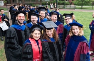 Graduates in their black gowns and caps. One in the front has a red stole. There are also two faculty members in the picture, wearing burgundy, black and blue gowns with blue caps